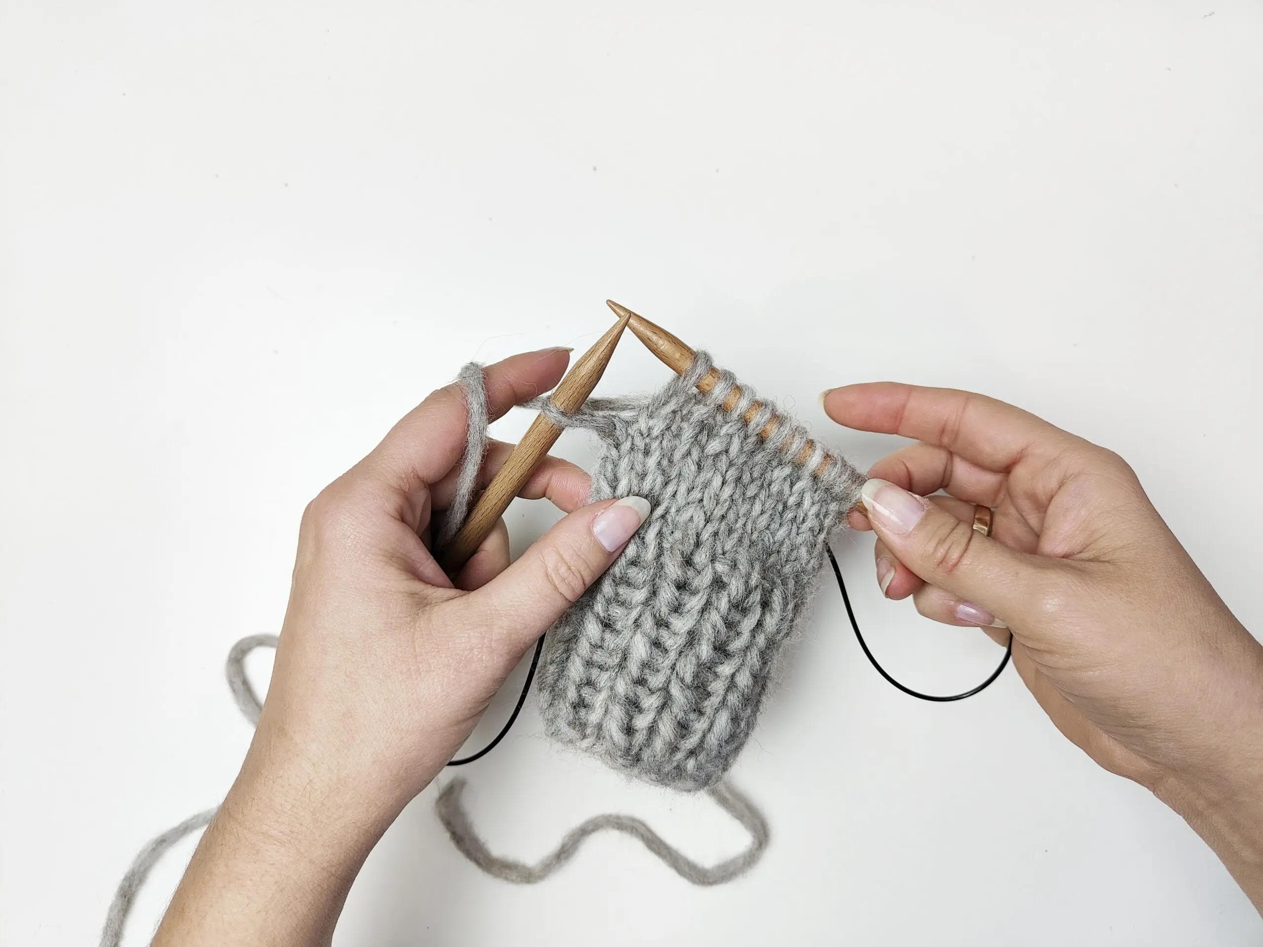 Zdjęcie przedstawia dłonie osoby, która jest w trakcie robienia na drutach. Dłonie trzymają drewniane druty i pracują z szarą włóczką. Lewa ręka trzyma drut, na który nawinięta jest włóczka, a prawa ręka trzyma drugi drut włożony w robótkę. Włóczka jest połączona z drutami na żyłce.