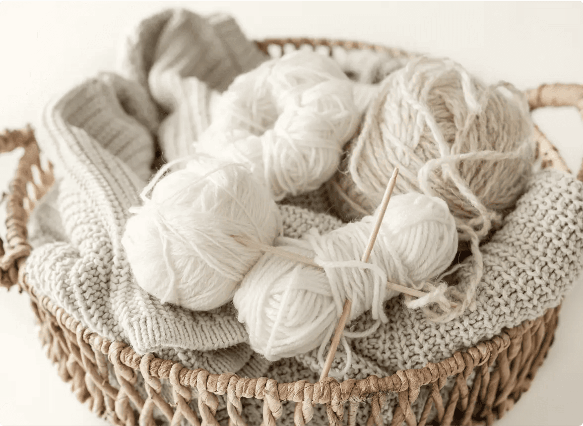 Na zdjęciu znajduje się kosz wypełniony włóczką i dzianiną. W koszu można zauważyć kilka kłębków włóczki w odcieniach bieli i beżu, a także druty do robienia na drutach. Włóczka jest ułożona na dzianinowych swetrach lub szalikach w podobnych kolorach.