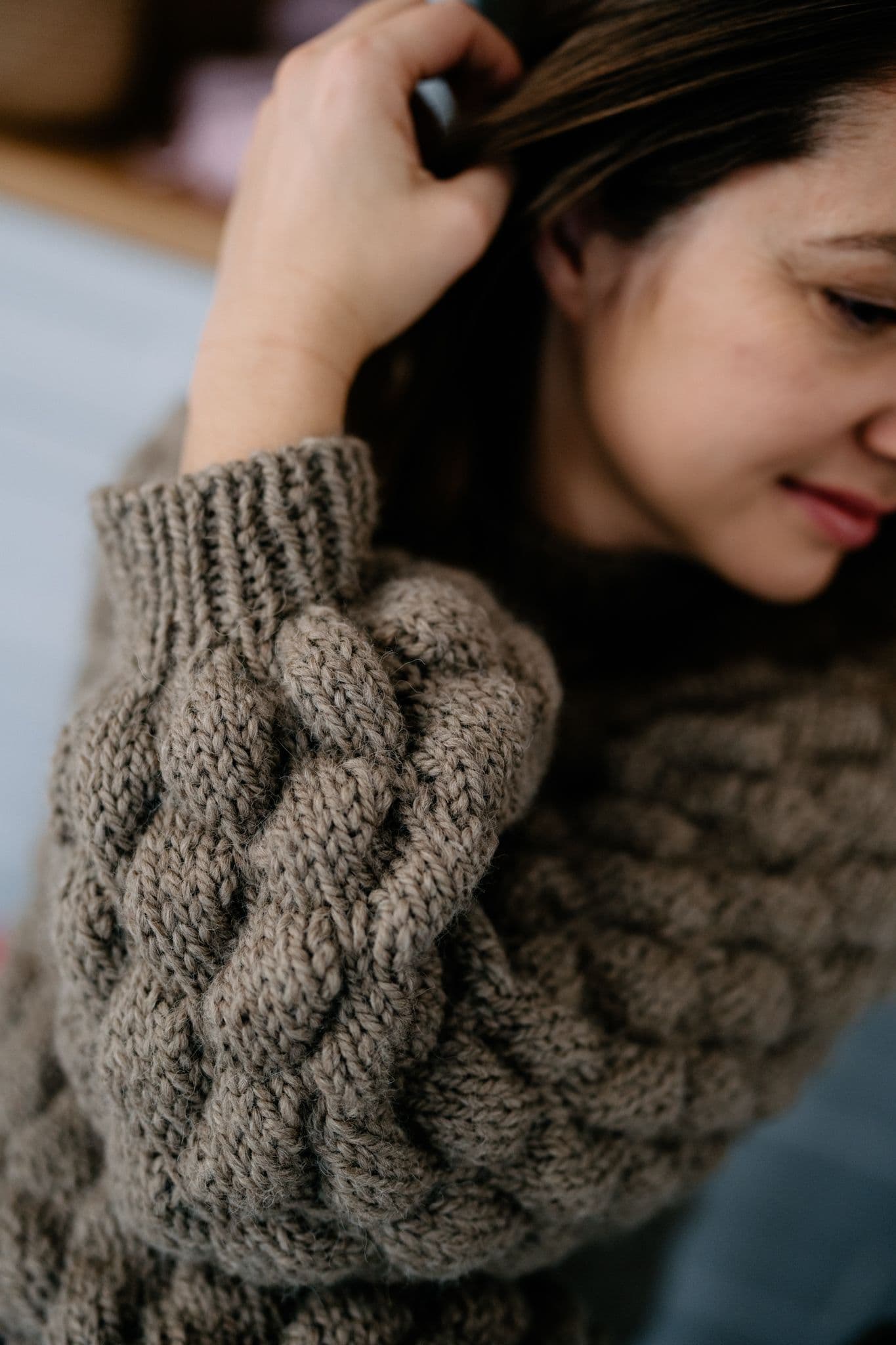 Obraz przedstawia zbliżenie na ramię i rękę osoby noszącej gruby, dziergany sweter o skomplikowanym wzorze warkocza. Sweter ma neutralny, ziemisty kolor, prawdopodobnie brązowy lub taupe. Osoba trzyma swoją rękę podniesioną do głowy, dotykając swoich włosów. Głównym punktem zainteresowania na obrazie jest tekstura i wzór swetra, co podkreśla kunszt wykonania dzianiny.