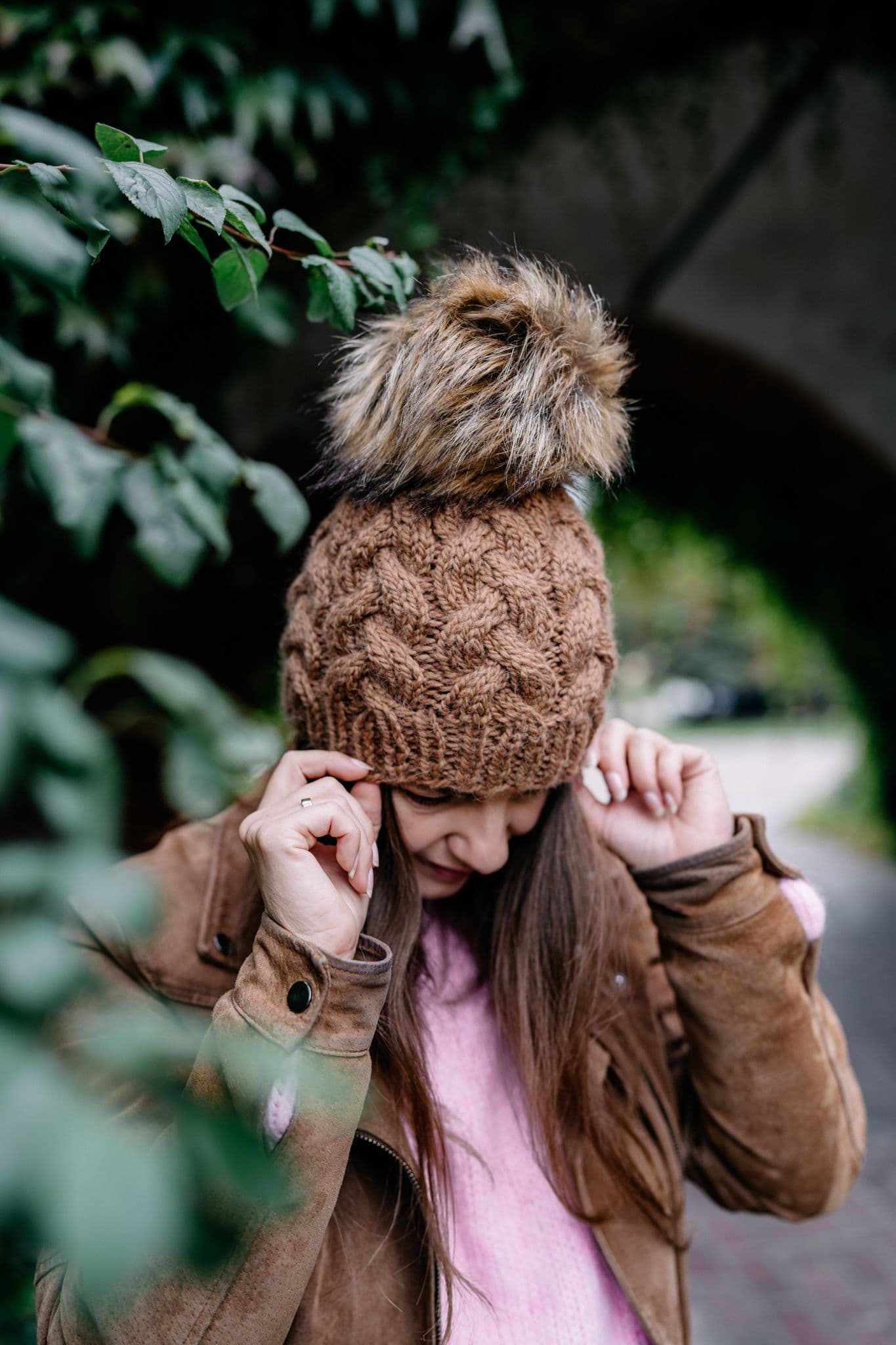 Obraz przedstawia kobietę, która ma na sobie wydzierganą, brązową czapkę z dużym puszystym pomponem oraz brązową kurtkę i różową bluzkę. Kobieta ma pochyloną głowę w dół, a dłońmi trzyma czapkę.