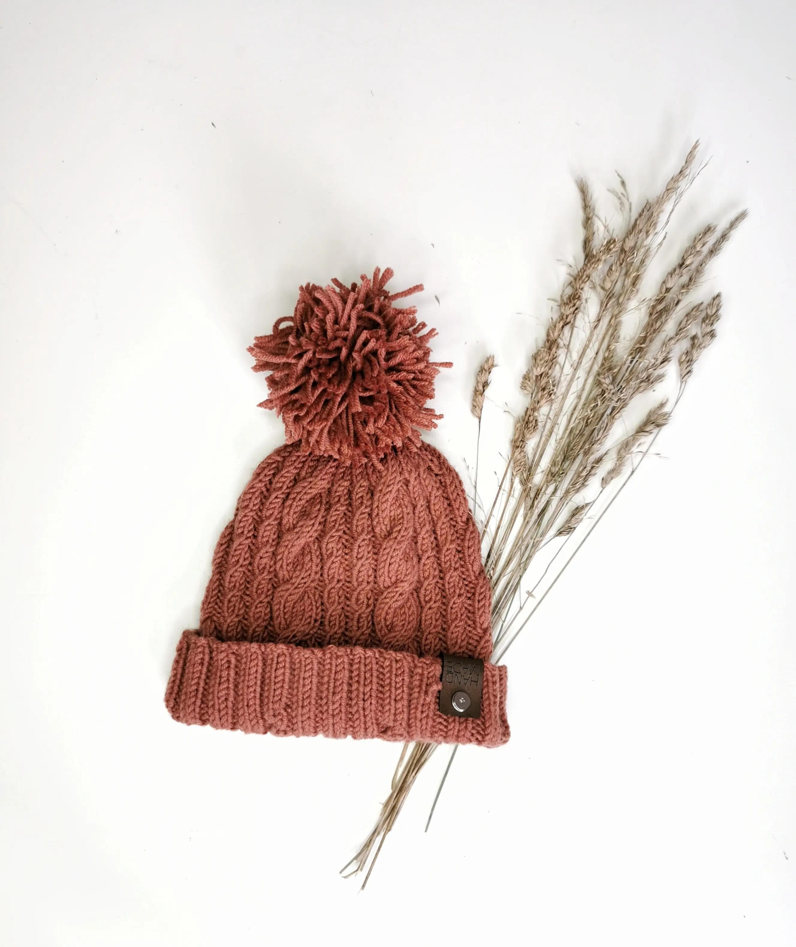 Obraz przedstawia dzierganą czapkę w kolorze czerwonawo-brązowym z dużym pomponem na górze. Czapka ma wzór warkoczy oraz wywinięty brzeg, na którym znajduje się mała skórzana naszywka z guzikiem i tekstem. Obok czapki leżą suszone trawy ułożone na białym tle.