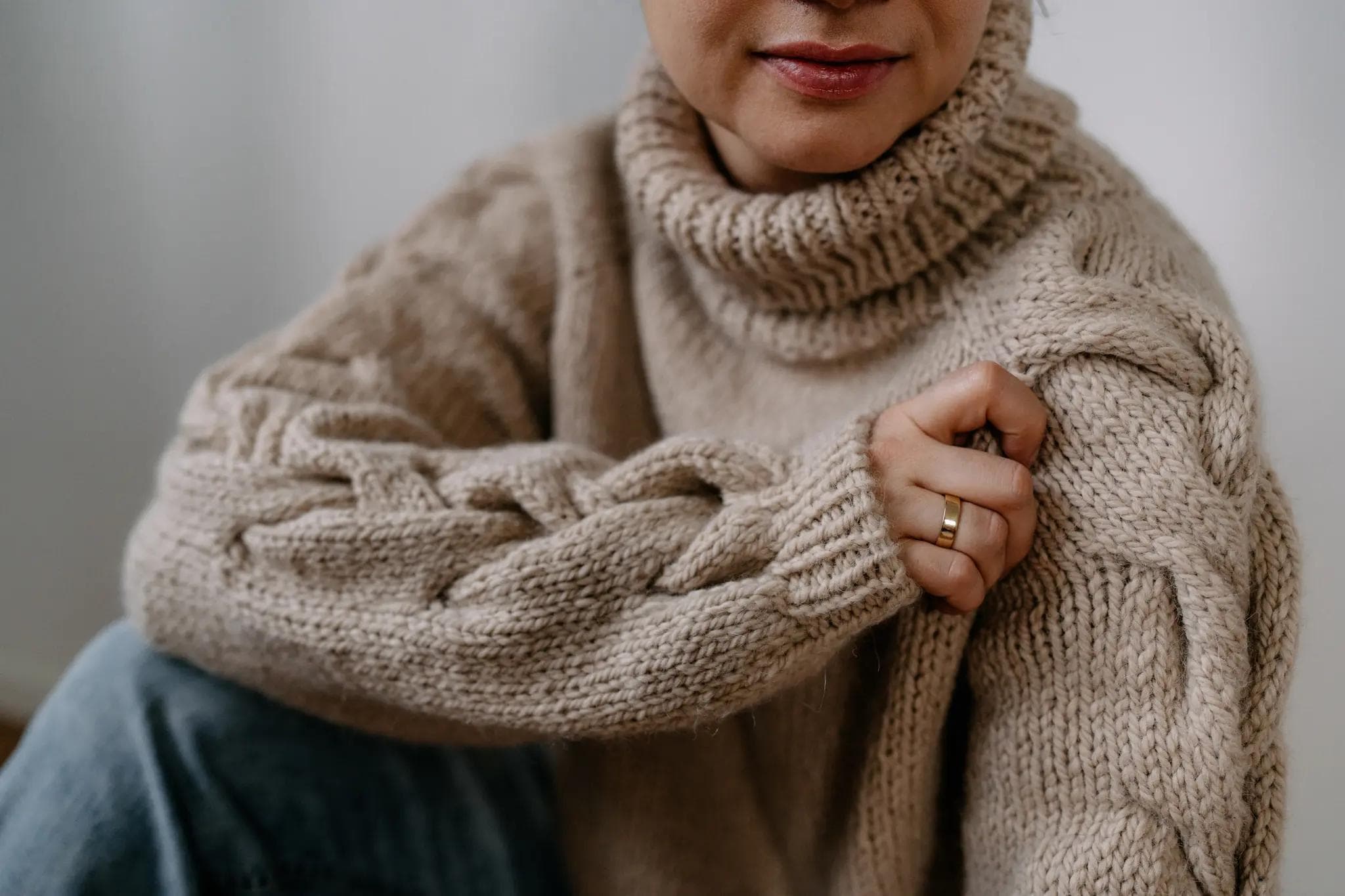 Na zdjęciu widoczna jest osoba ubrana w gruby, beżowy sweter z wysokim golfem i warkoczowym wzorem na rękawie. Sweter wykonany jest z puszystej, mięsistej włóczki, co nadaje mu przytulny wygląd i teksturę. Osoba siedzi, lekko przyciągając rękaw do siebie, eksponując splot na rękawie i pierścionek na palcu. Tło jest neutralne, co dodatkowo podkreśla ciepły odcień swetra i jego strukturalny wzór.