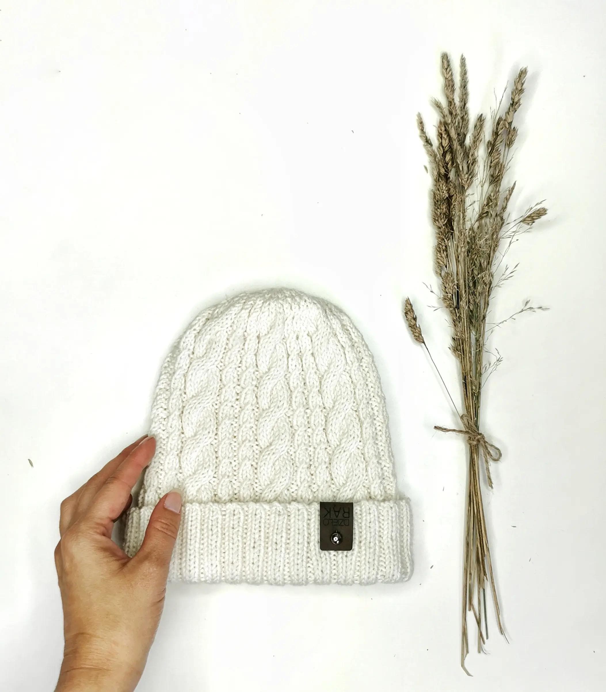 Na zdjęciu znajduje się biała, dziergana czapka z grubym splotem, trzymana przez rękę. Obok czapki, po prawej stronie, znajduje się bukiet suchych traw związanych sznurkiem.