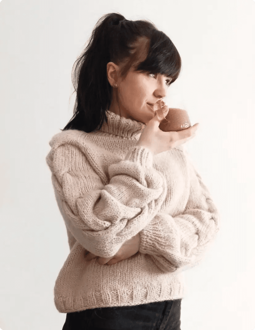 Na zdjęciu widzimy Martę w jasnym, grubym swetrze o warkoczowym splocie, która trzyma filiżankę w dłoniach, przybliżając ją do ust, jakby delektowała się jego zapachem. Ma ciemne, długie włosy związane w kucyk, a jej grzywka opada na czoło. Jej wyraz twarzy sugeruje chwilę zadumy lub relaksu. Kobieta stoi na jasnym, jednolitym tle.