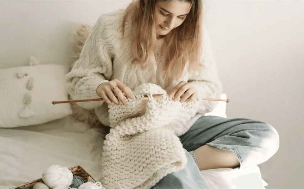 Obraz przedstawia kobietę siedzącą na łóżku lub kanapie i zajmującą się robótką na drutach. Kobieta ma na sobie przytulny, kremowy sweter i jasnoniebieskie dżinsy. Projekt dziewiarski wydaje się być dużym, grubym kawałkiem, prawdopodobnie kocem lub szalikiem, wykonanym z grubej, kremowej włóczki. Kobieta używa drewnianych drutów do robienia na drutach. Obok kobiety znajduje się koszyk z kilkoma kłębkami włóczki w różnych kolorach, w tym białym i szarym. W tle widoczna jest poduszka z dekoracyjnymi elementami.