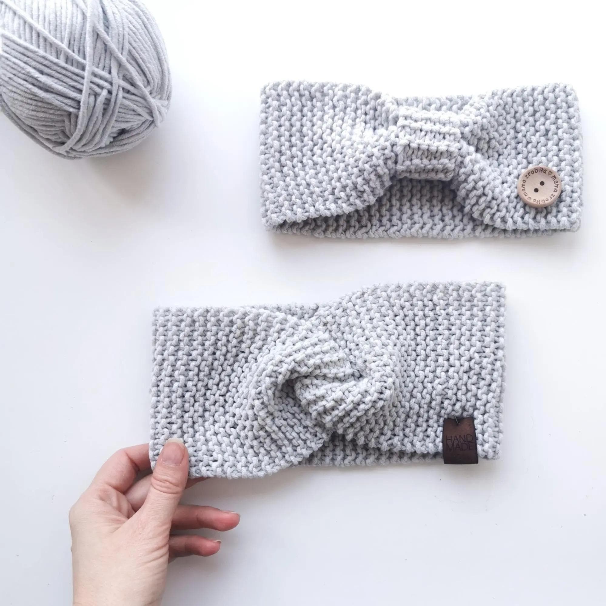 Obraz przedstawia dwa dziergane opaski i kłębek włóczki. Opaski są jasnoszare i mają różne wzory. Górna opaska ma kształt kokardy, z guzikiem po prawej stronie. Dolna opaska, którą trzyma ręka, ma skręcony węzeł na środku i małą metkę z napisem „HAND MADE” z jednej strony. Kłębek włóczki jest umieszczony w górnym lewym rogu obrazu.