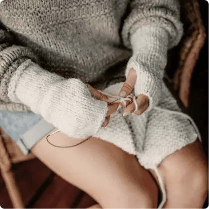 Na zdjęciu kobieta w beżowym sweterku i jasnych spodenkach z jasnymi mitenkami na dłoniach. Kobieta w dłoniach trzyma druty i jasną włóczkę, a na kolanach leży najprawdopodobniej robiony przez nią szalik.