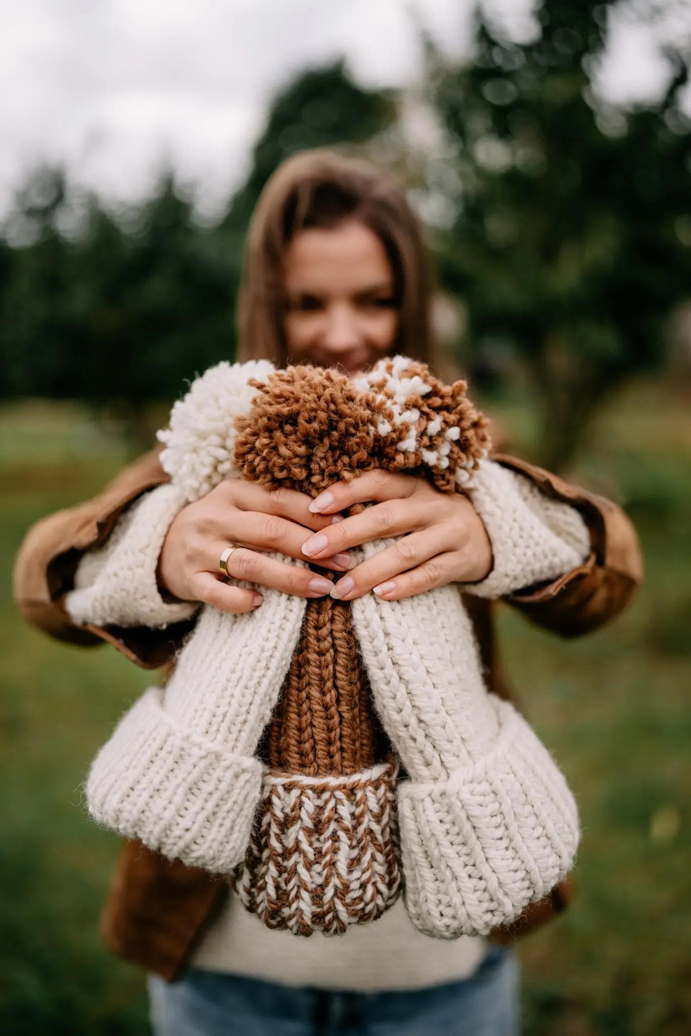 Obraz przedstawia uśmiechniętą kobietę, która trzyma w wyciągniętych rękach trzy wydziergane czapki. Czapki są kolorach bieli i brązu z pomponami. Kobieta ubrana jest w brązową kurtkę, jasny sweterek i niebieskie dżinsy.