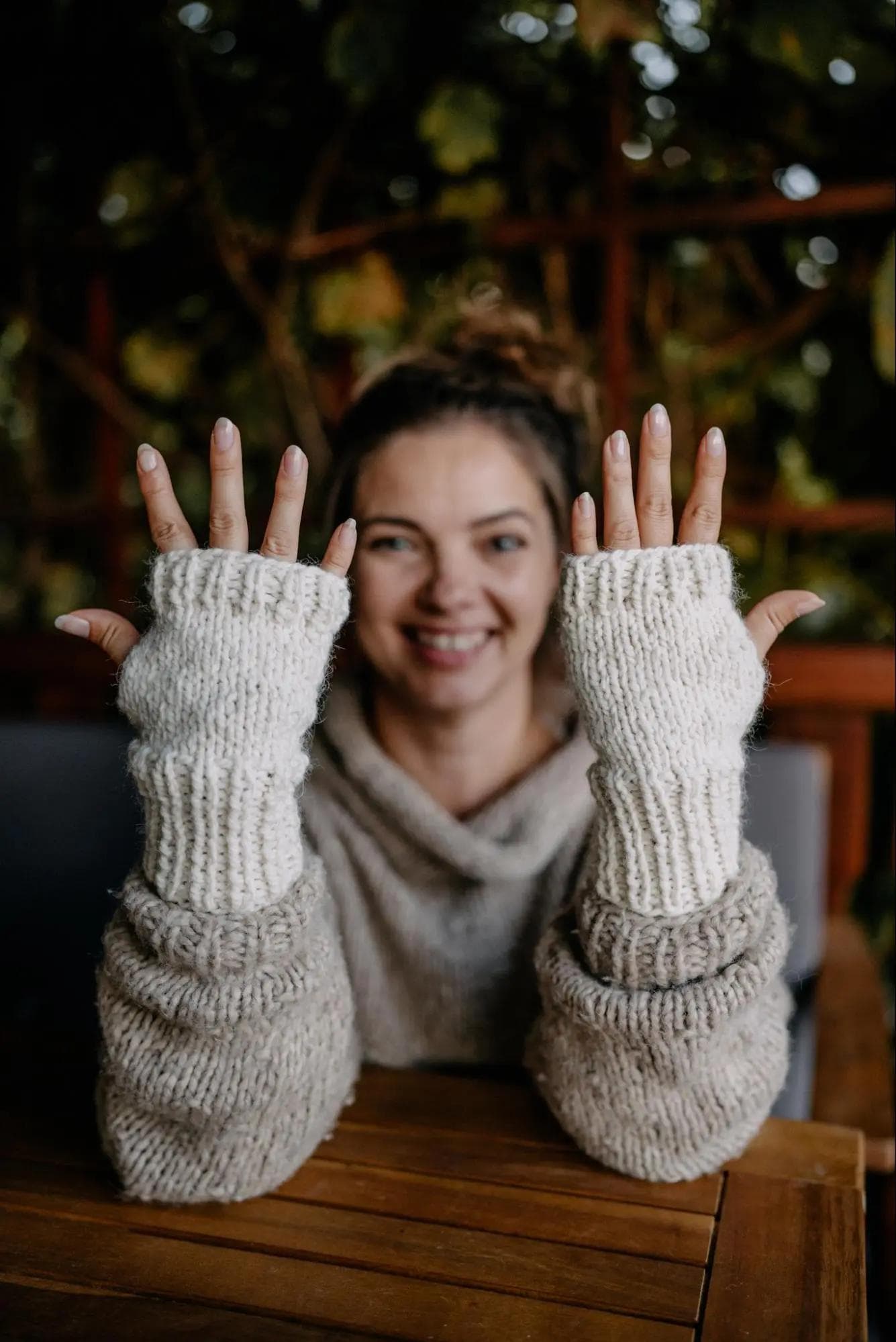 Na zdjęciu uśmiechnięta kobieta, która ma zamazaną twarz. Kobieta ma na sobie jasny, dziergany sweter i pasujące do niego rękawiczki bez palców. Osoba trzyma obie ręce w górze, prezentując rękawiczki. Tło jest rozmyte, a w tle widać trochę zieleni.