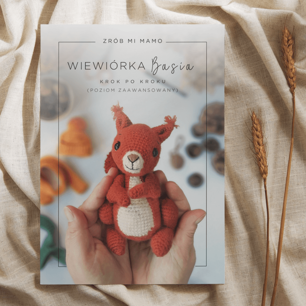 Na zdjęciu widzimy książkę zatytułowaną “Zrób mi, Mamo: Wiewiórka Basia krok po kroku (poziom zaawansowany)”. Jest to przewodnik po tworzeniu zabawek na szydełku, w tym przypadku wiewiórki. Na okładce znajduje się zdjęcie dwóch rąk trzymających gotową, szydełkowaną wiewiórkę. W tle widoczne są miękkie tkaniny i kłosy zboża.