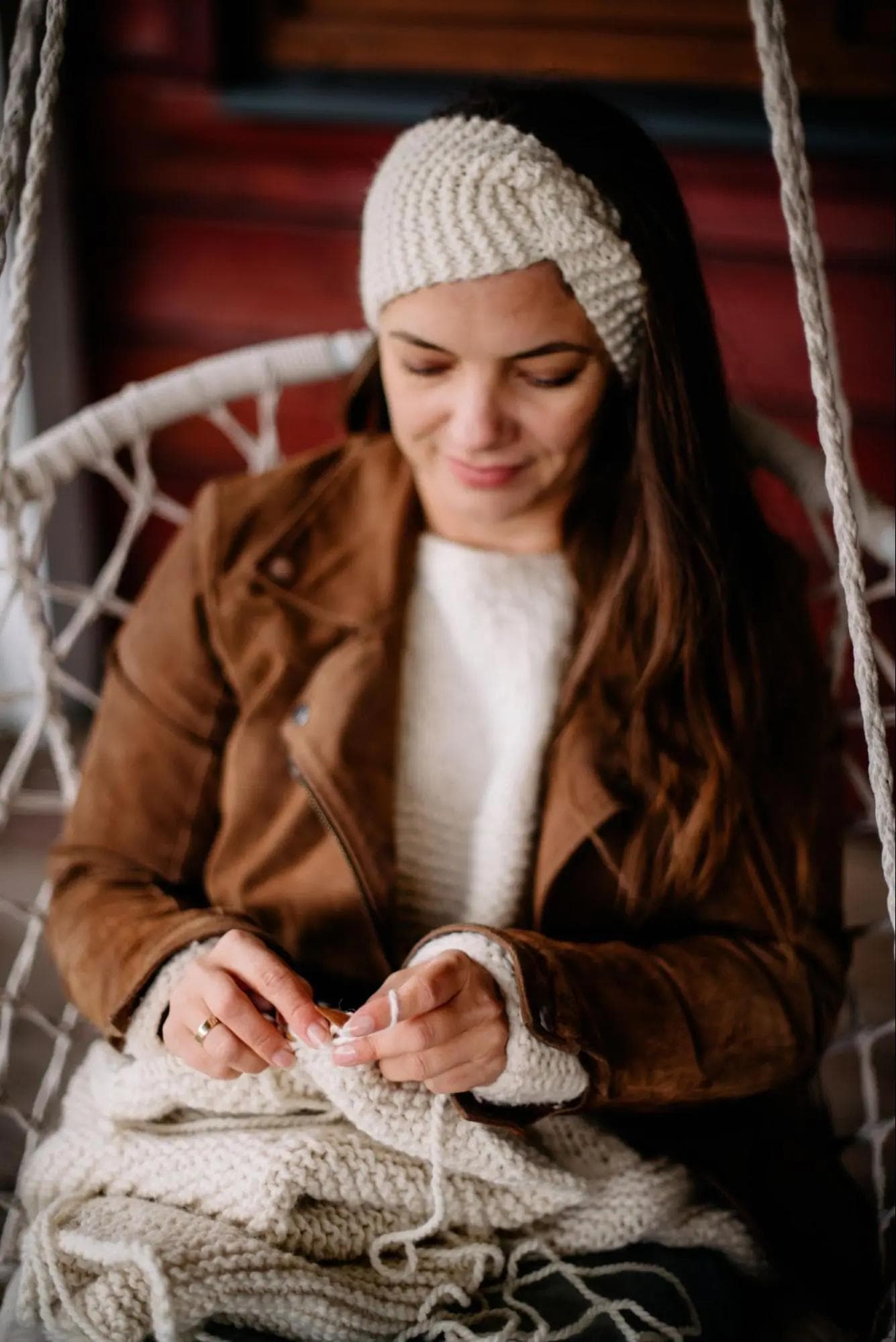 Obraz przedstawia skupioną kobietę, która siedzi na huśtawce i dzierga. Kobieta ma na sobie brązową kurtkę, biały sweter i białą opaskę na głowie. W dłoniach trzyma druty i pracuje nad kawałkiem białej dzianiny. Tło to drewniana konstrukcja, prawdopodobnie weranda lub chatka.