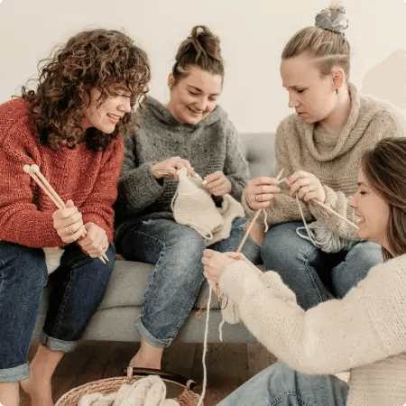 Na zdjęciu widzimy cztery kobiety, z których trzy siedzą na szarej kanapie. Kobiety ubrane są w różne kolory swetry i dżinsy. Każda z nich trzyma druty, a na podłodze przed nimi znajduje się koszyk z włóczką.