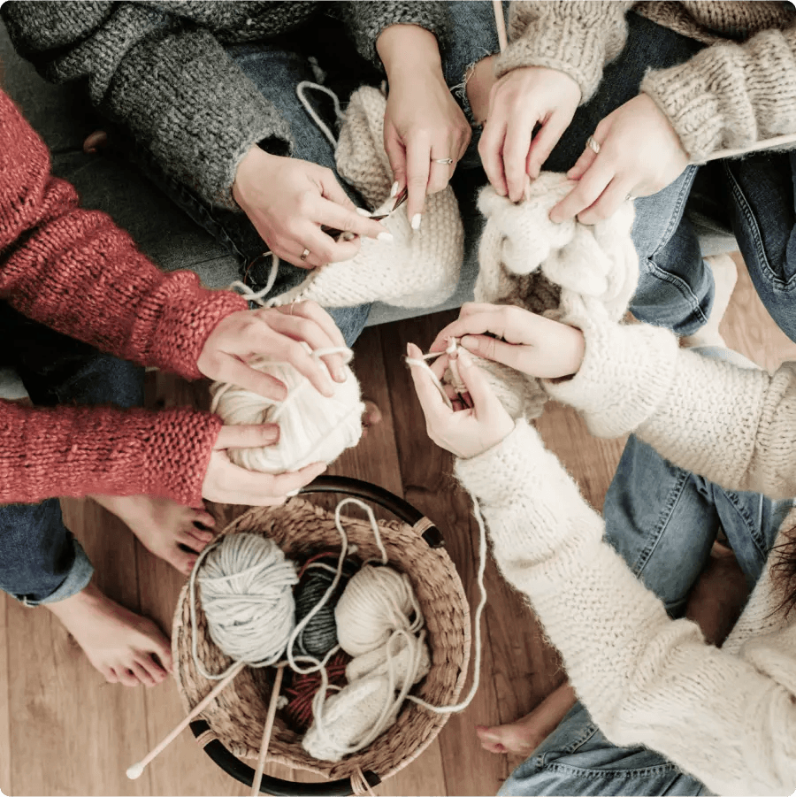 Obraz przedstawia grupę osób siedzących razem i zajmujących się robieniem na drutach. Każda z nich nosi przytulny sweter, a ich ręce są zajęte drutami i włóczką w różnych kolorach, zebranymi w koszyku na podłodze. Scena sugeruje wspólne, relaksujące zajęcie, prawdopodobnie spotkanie robótek ręcznych lub luźne zgromadzenie przyjaciół, którzy lubią razem robić na drutach.