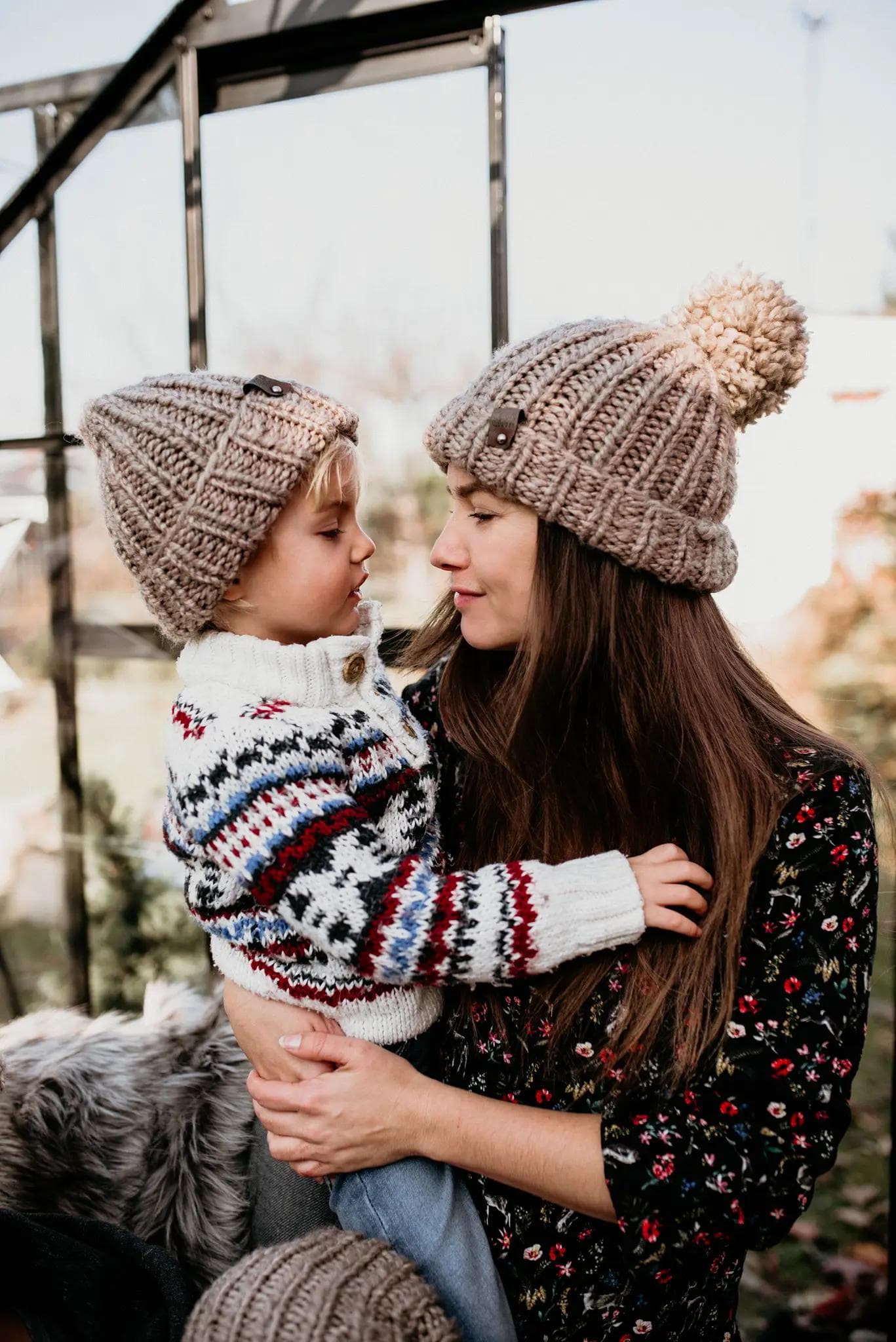 Zdjęcie przedstawia matkę trzymającą swojego syna, którzy patrzą na siebie. Oboje mają na sobie beżowe zimowe czapki. Chłopiec jest ubrany w wełniany, biały sweter we wzory, a kobieta w ciemną bluzkę w kwiaty. Tło zdjęcia jest rozmyte.